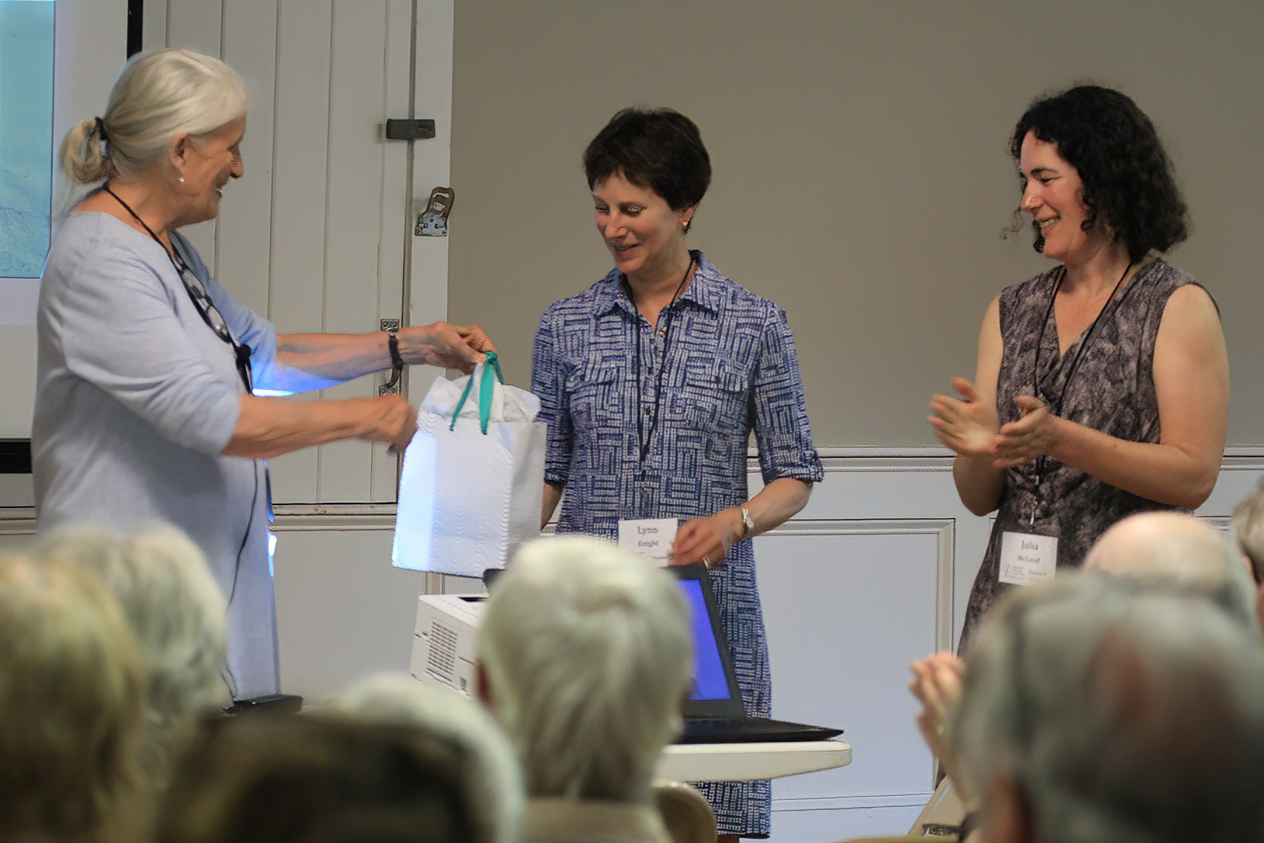 two women present a third woman with a bagged gift in front of crowd