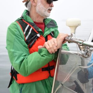 older man in green jacket and orange life vest stands at helm of boat.