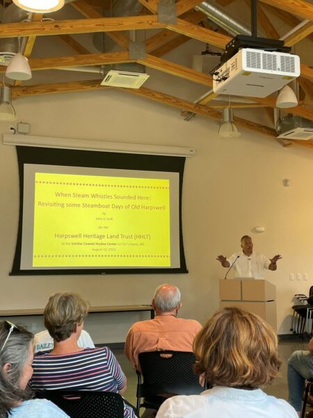 John Goff stands speaking at podium next to yellow slide presentation