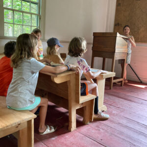 child stands at podium while peers sit at old fashioned desks in schoolhouse