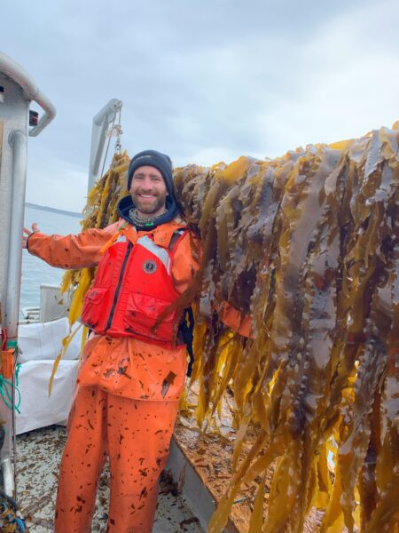 man stands on boat smiling next to line full of kelp