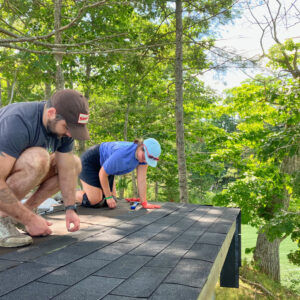 two people kneel on shed roof while adding new shingles