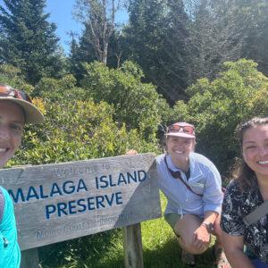 three people stand and smile with the sign for Malaga Island Preserve