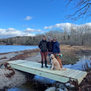three women and a dog stand on a small bridge over a stream