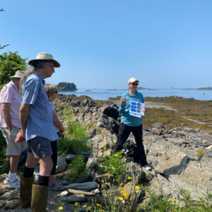 people stand around rocky shoreline and face woman presenting diagrams