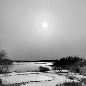 black and white photo of sun over small pond with ocean beyond.